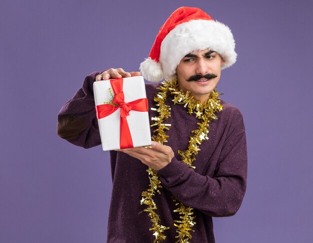 Young mustachioed man wearing christmas santa hat with tinsel around his neck holding christmas present looking confused and displeased  standing over purple background