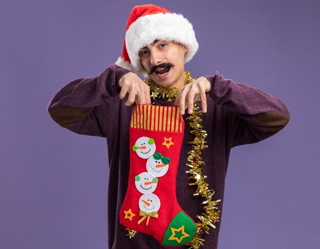 Young mustachioed man wearing christmas santa hat with tinsel around his neck holding christmas stocking looking at camera surprised and happy standing over purple background