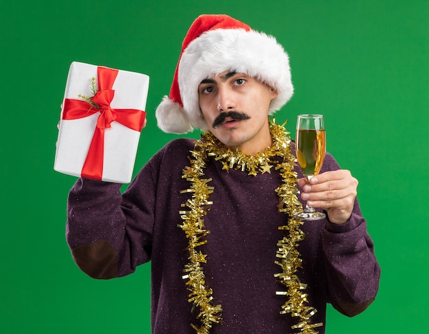 緑の背景の上に立って混乱しているカメラを見てシャンパンとクリスマスプレゼントのガラスを保持している彼の首に見掛け倒しのクリスマスサンタ帽子をかぶっている若い口ひげを生やした男