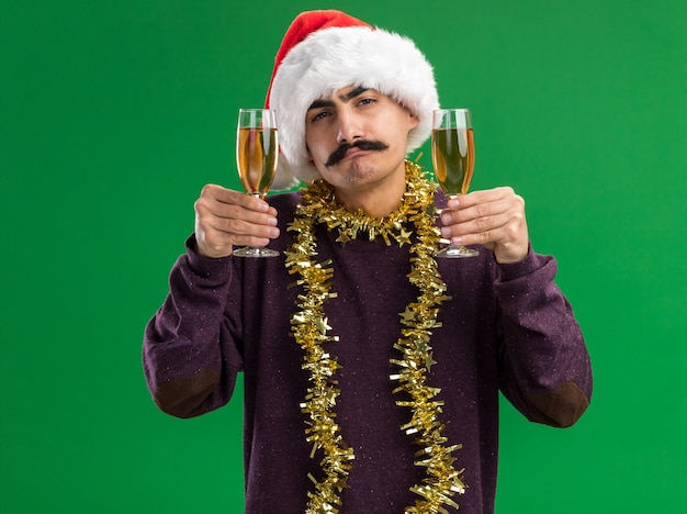 녹색 배경 위에 서있는 자신감있는 표정으로 카메라를보고 샴페인 두 잔을 들고 그의 목에 반짝이와 크리스마스 산타 모자를 쓰고 젊은 mustachioed 남자