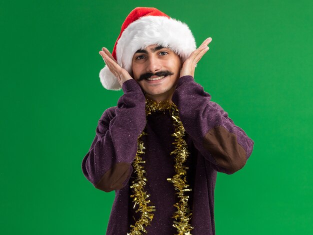 Бесплатное фото Молодой усатый мужчина в рождественской шапке санта-клауса с мишурой на шее, глядя в камеру со счастливым лицом, весело улыбаясь, стоя на зеленом фоне