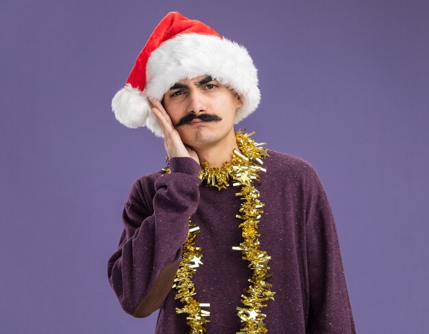 Young mustachioed man wearing christmas santa hat with tinsel around his neck looking at camera confused with hand on his face  standing over purple background