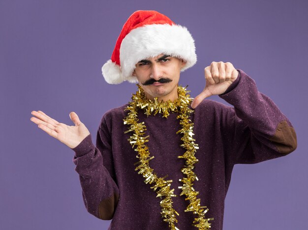 Молодой усатый мужчина в рождественской шапке санта-клауса с мишурой на шее, недовольно смотрящий в камеру, показывает большие пальцы вниз и представляет руку, стоящую на фиолетовом фоне