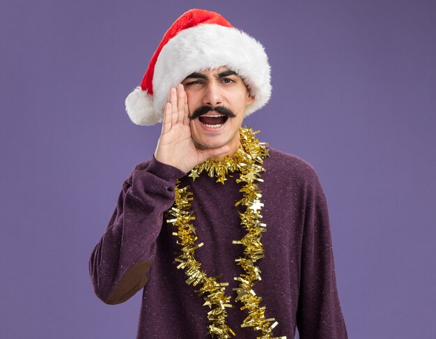 young mustachioed man wearing christmas santa hat with tinsel around his neck shouting with hand near mouth standing over purple background