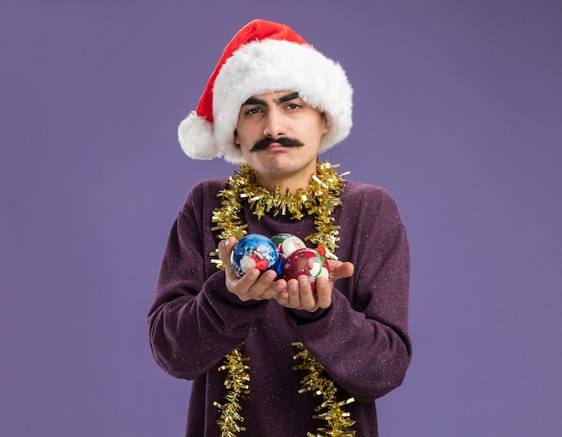 Free photo young mustachioed man wearing christmas santa hat with tinsel around his neck showing christmas balls  confused  standing over purple wall