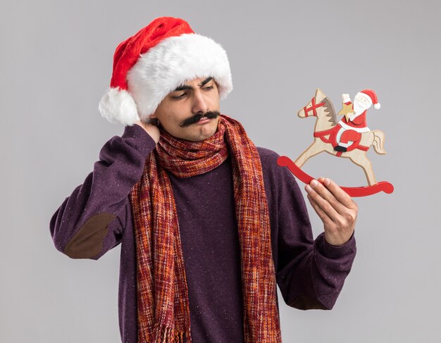 молодой усатый мужчина в рождественской шапке санта-клауса с теплым шарфом на шее, держащий рождественскую игрушку, смотрит на нее смущенно и очень взволнованно, стоя на белом фоне