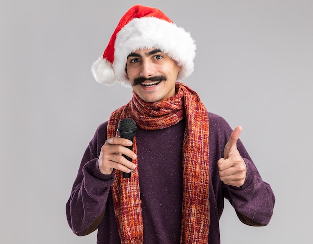 Young mustachioed man wearing christmas santa hat with warm scarf around his neck holding microphone looking smiling cheerfully showing thumbs up 