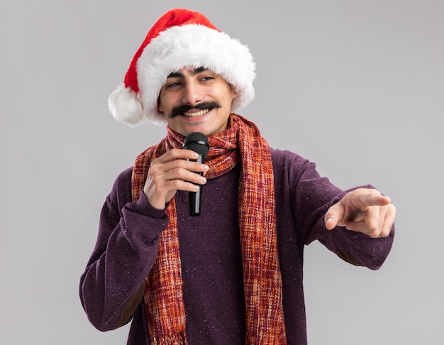 Молодой усатый мужчина в рождественской шапке санта-клауса с теплым шарфом на шее держит микрофон, улыбаясь, указывая указательным пальцем в сторону, стоя над белой стеной