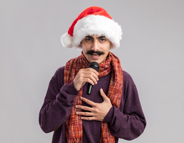 Young mustachioed man wearing christmas santa hat with warm scarf around his neck speaking to microphone looking with smile on face 