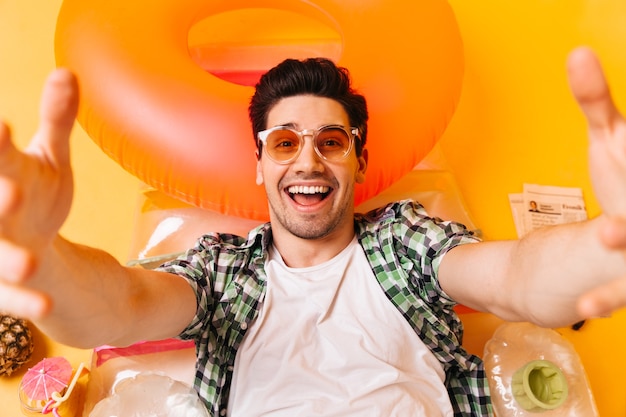 Young naughty man in plaid shirt and orange glasses smiles and takes selfie on inflatable mattress.