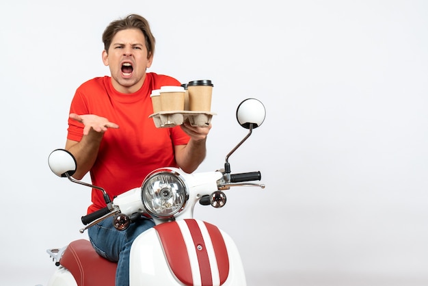 Uomo giovane corriere nervoso in uniforme rossa che si siede sullo scooter e che tiene gli ordini sulla parete gialla