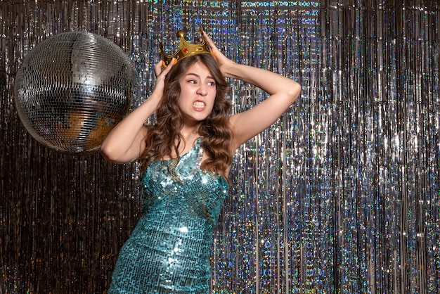 Young nervous unsatisfied beautiful lady wearing blue green shiny dress with sequins with crown in the party