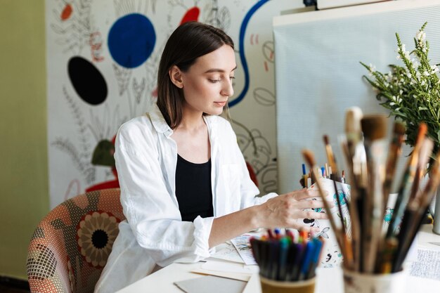 Giovane bella ragazza alla scrivania che sceglie sognante l'immagine mentre trascorre del tempo nel posto di lavoro creativo a casa