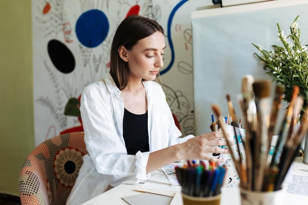 Young nice girl at the desk dreamily choosing picture while spending time in creative workplace at home