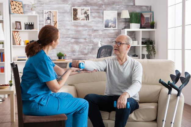 Young nurse doing physiotherapy treatment in nursing home with senior man using dumbbells.