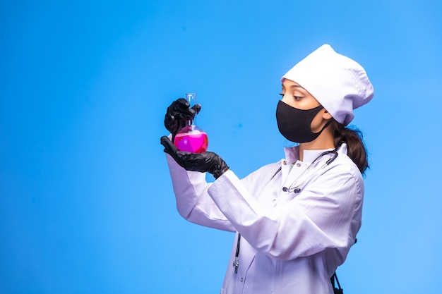 顔と手のマスクの若い看護師がテストフラスコを保持し、青い壁に化学反応を追う
