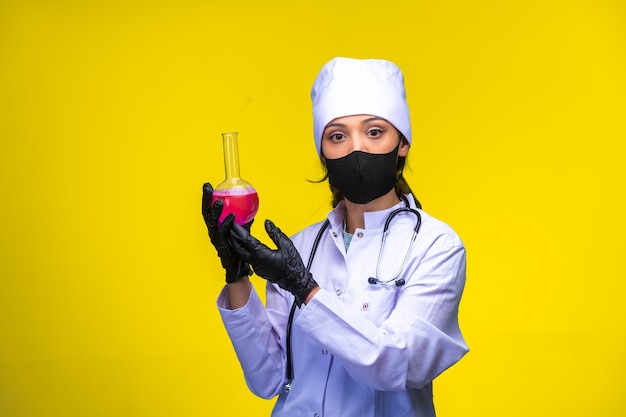 Young nurse in face and hand mask holds a test flask with pink liquid.