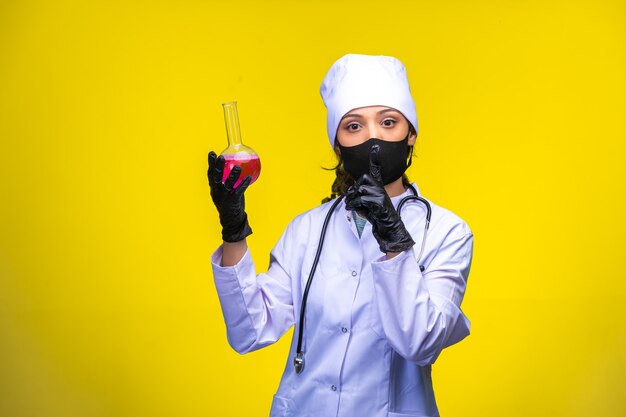 Young nurse in face and hand mask holds a test flask on yellow.