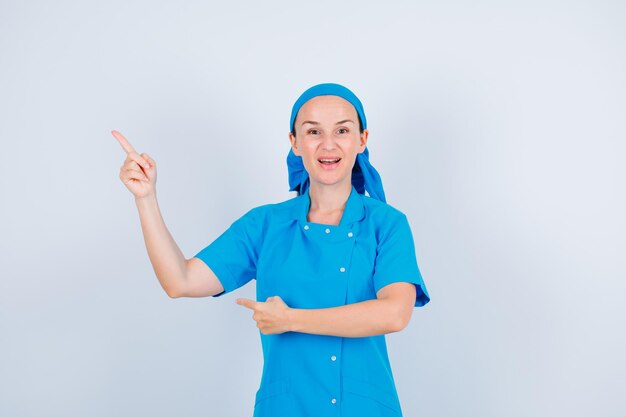 Young nurse is looking at camera by pointing left up with forefinger on white background