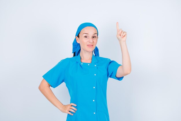 Young nurse is pointing up with forefinger and putting other hand on waist on white background