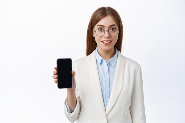 Young office woman showing smartphone screen, wearing business suit and glasses, smiling and looking professional at front, standing over white wall