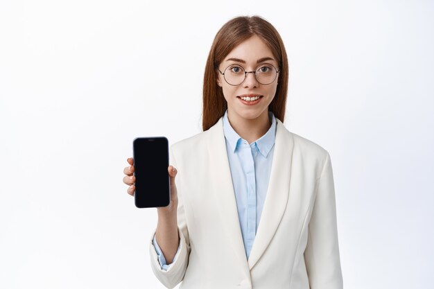 스마트폰 화면을 보여주는 젊은 사무실 여성, 비즈니스 정장과 안경을 쓰고 웃고, 정면에서 전문가를 바라보고, 흰 벽 위에 서 있다