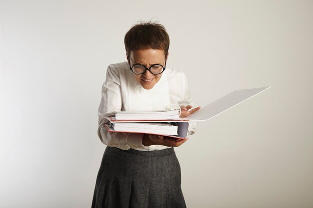 Young old-fashioned looking teacher in round black glasses squints in disbelief on page inside one of the two binders she holds isolated on white.