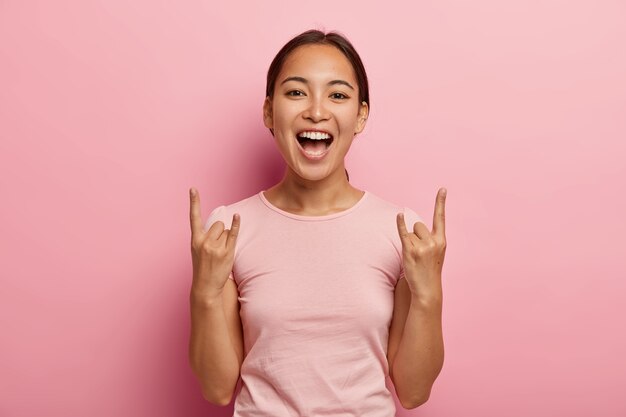 Young overjoyed brunette woman with Asian appearance, poses with raised arms horns and makes rock gesture, being upbeat and sartisfied, exclaims happily, wears casual pink t shirt, poses in 