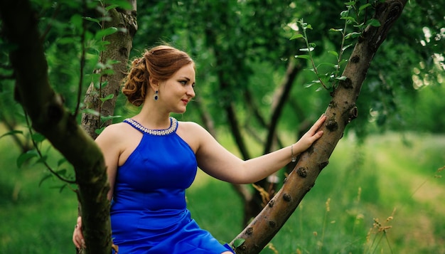 Молодая полная девушка в голубом платье позирует на фоне весеннего сада, сидя на дереве