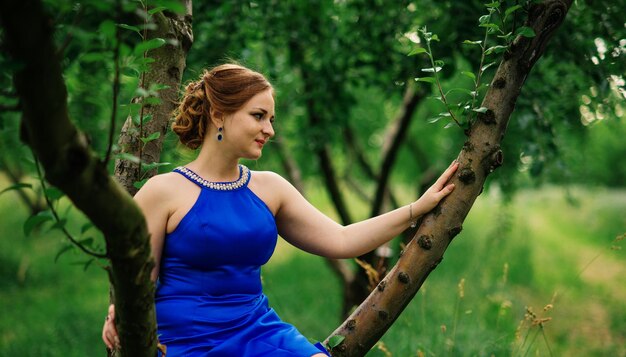 Молодая полная девушка в голубом платье позирует на фоне весеннего сада, сидя на дереве