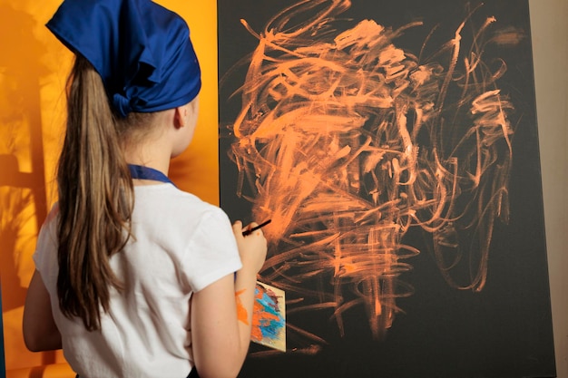 Young painter painting masterpiece with orange color, using colormix tray with aquarelle dye to paint artwork creation on canvas. Child using paintbrush with watercolor, artistic development.