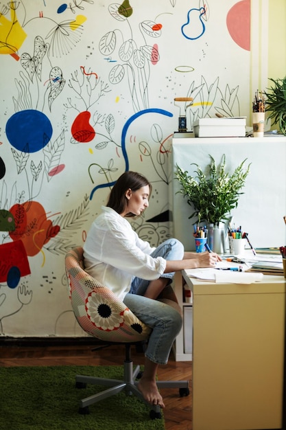 책상에 앉아 있는 흰 셔츠를 입은 젊은 화가는 집에서 배경에 큰 다채로운 패턴의 캔버스를 사용하여 신중하게 그림을 그립니다.