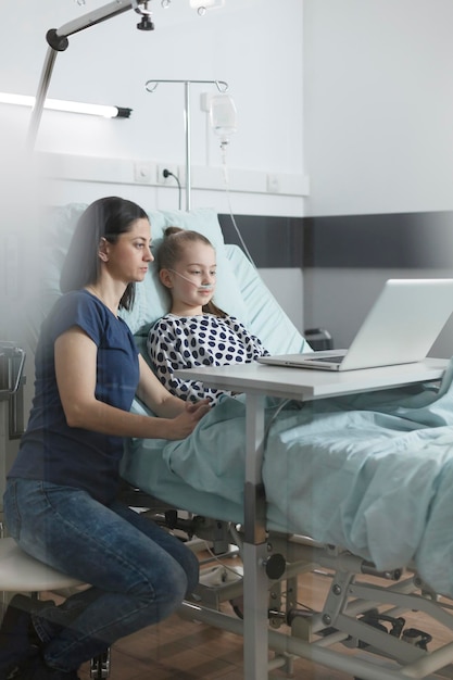 무료 사진 젊은 부모와 몸이 좋지 않은 어린 딸이 노트북에서 웹 페이지를 서핑합니다. 현대적인 컴퓨터를 사용하면서 편안한 엄마와 딸이 소아과 병동에 앉아 있습니다.
