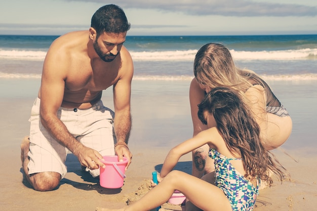 Young parents and cute little girl romping with wet sand on beach, digging with toy shovel, bucket and bowl
