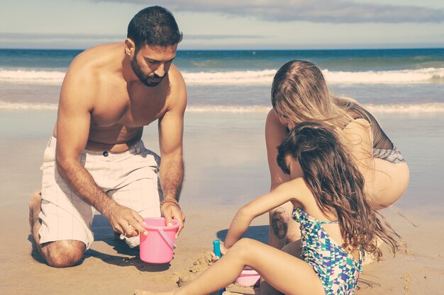 젊은 부모와 해변에 젖은 모래로 뛰어 다니는 귀여운 소녀, 장난감 삽, 양동이와 그릇으로 파고