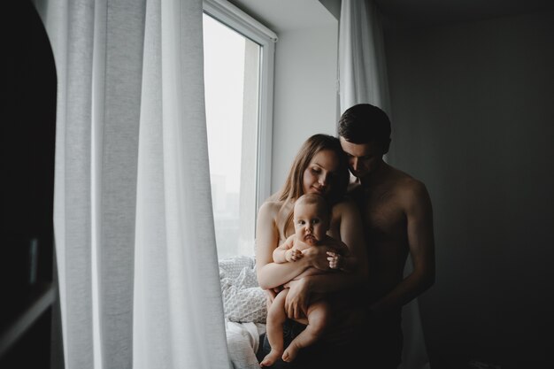 Young parents hold newborn baby standing before a bright window in a dark room