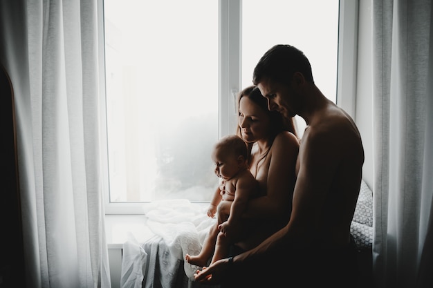 Young parents hold newborn baby standing before a bright window in a dark room