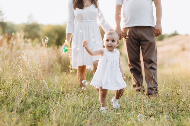 Young parents walk with their lovely little daughter in jeans dress across the field