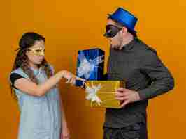 Free photo young party coulpe wearing masquerade eye mask girl putting finger on gift box in guy hand isolated on orange