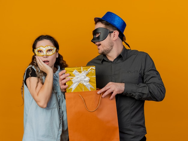 Young party couple wearing masquerade eye mask guy given gift at surprised girl isolated on orange