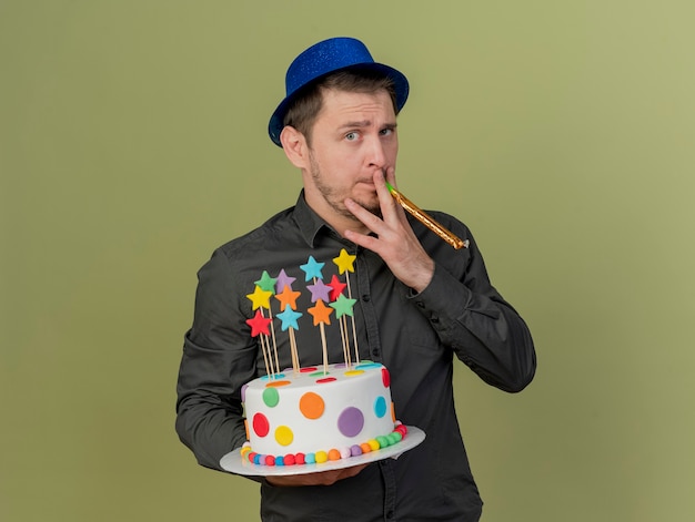 young party guy wearing black shirt and blue hat holding cake and blowing party blower isolated on olive green