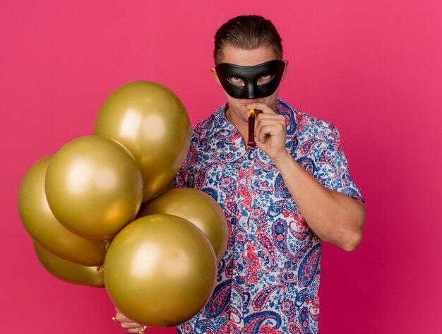 Young party guy wearing masquerade eye mask holding balloons and blowing party blower isolated on pink