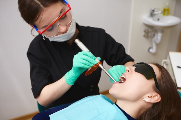 치과 치료 빛을 사용하여 여성 위생사가 치료하는 그녀의 이빨을 받고 검은 고글에 젊은 환자