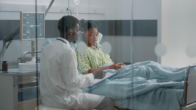 Young patient receiving medical consultation from doctor in hospital ward. Medical specialist talking to ill woman in bed about prescription treatment and healthcare to heal disease.