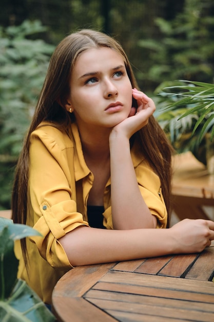 Young pensive brown haired woman in yellow shirt leaning head on hand thoughtfully looking up around green leaves in city park