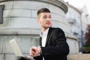 Free photo young pensive businessman in classic black suit with wireless earphones dreamily looking aside with hand watch and laptop on knees outdoor