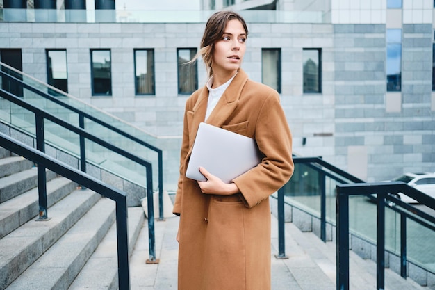 Foto gratuita giovane donna di affari casual pensierosa in cappotto con il computer portatile che distoglie lo sguardo sognante all'aperto