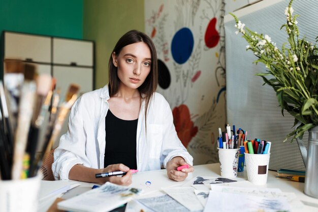 Young pensive girl sitting at the desk dreamily looking in camera with pink marker in hand at home