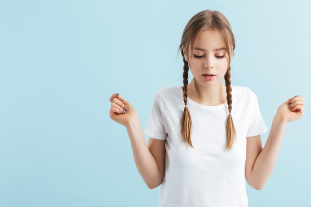 Young pensive girl with two braids in white tshirt thoughtfully looking down over blue background isolated