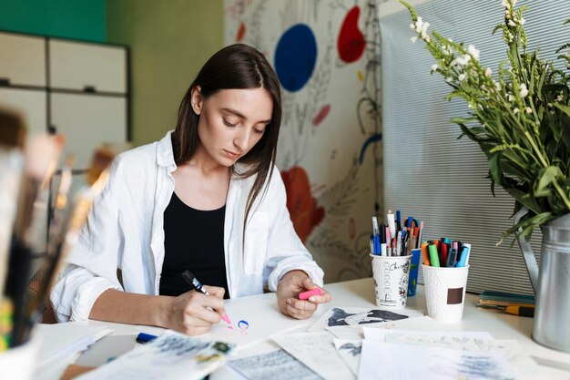 Молодой задумчивый художник за столом мечтательно рисует розовым маркером, проводя время на творческом рабочем месте дома